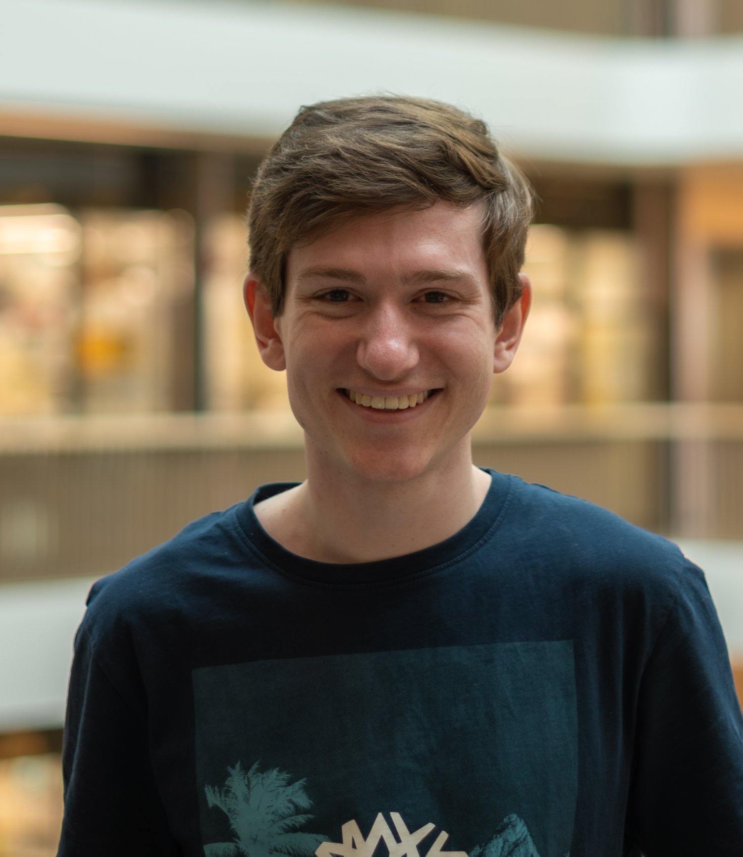 Person i mörk T‑shirt med tryck står inomhus framför en suddig bakgrund av ett modernt atrium.