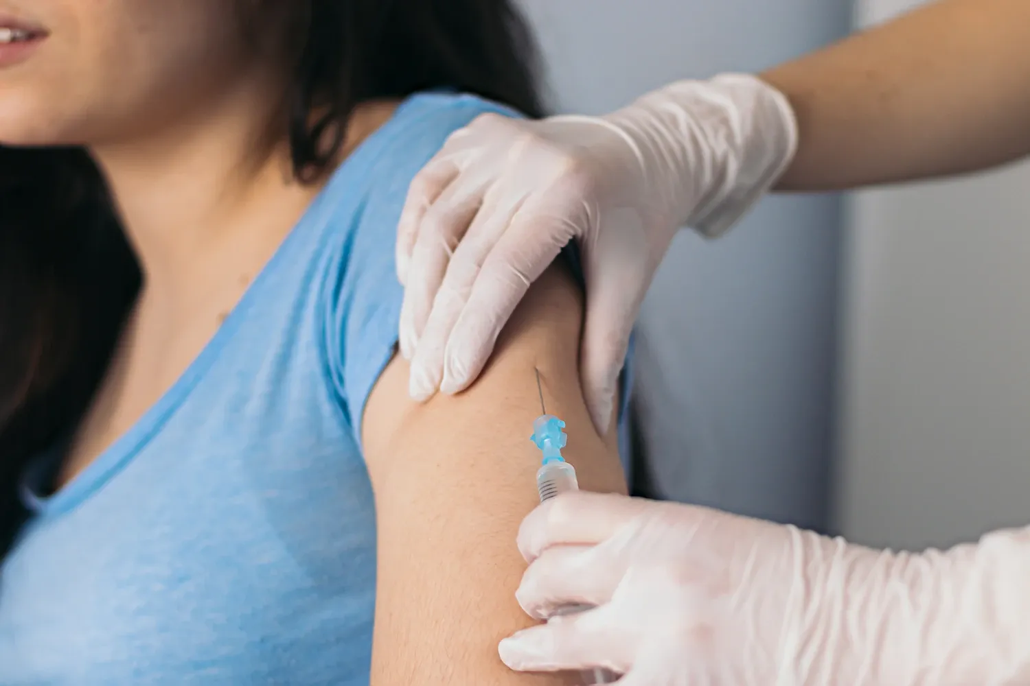 En närbild av en vaccination som ges i överarmen. En person i blå tröja får en injektion medan vårdpersonal med handskar håller armen och för in sprutan.