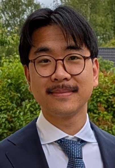 Mörkhårig man med glasögon och mustasch iklädd blå kavaj, vit skjorta och blårutig slips.