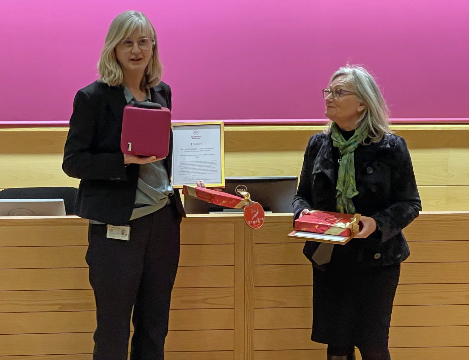 Maria Ankarcrona, prefekt och Carin Ullman, administrativ chef, har precis mottagit KI:s arbetsmiljöpris 2024. De står vid scenen i rum Berzelius på Campus Solna.