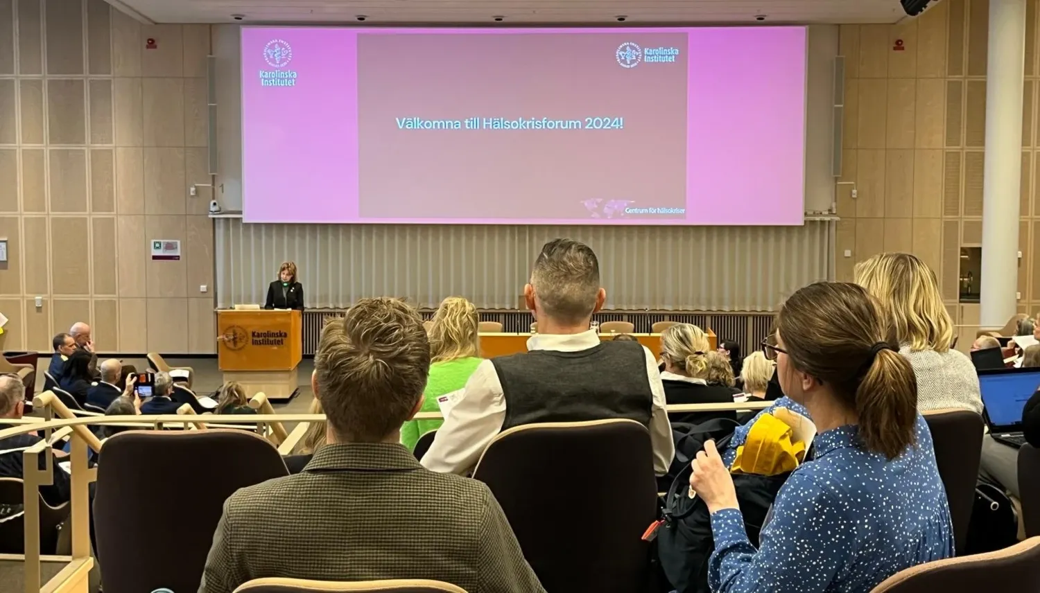 En kvinna står i en talarstol framför en skärm där det står Hälsokrisforum 2024. I förgrunden syns en publik