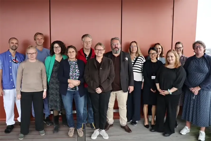 Gruppbild av 14 personer vid en brunröd vägg