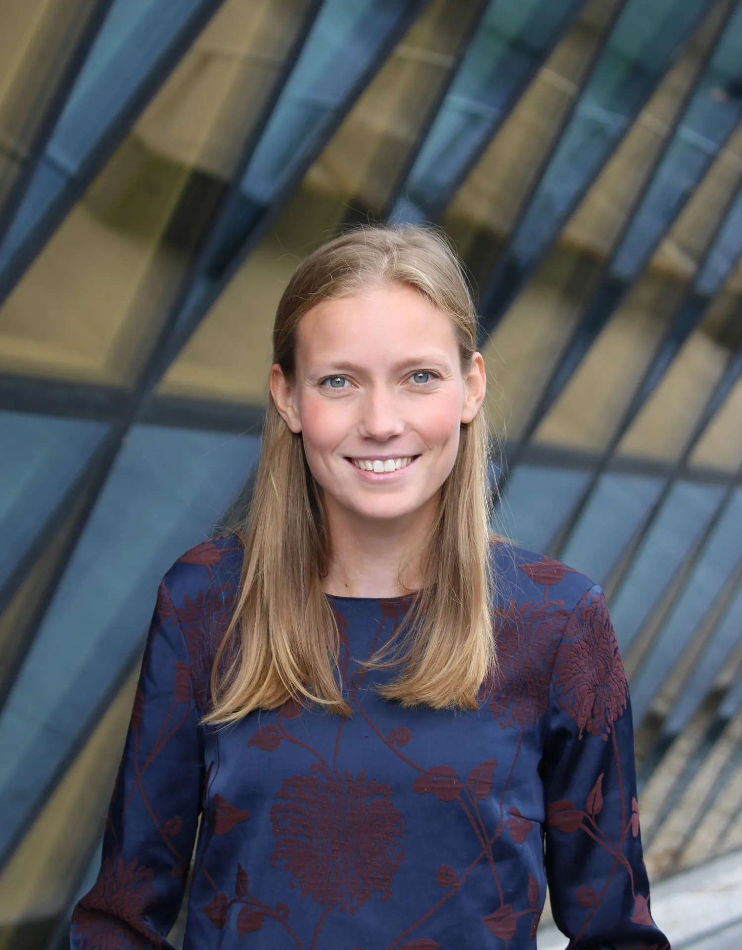 Kvinna med blont hår klädd i en blå topp står framför en byggnad med glasfasad.