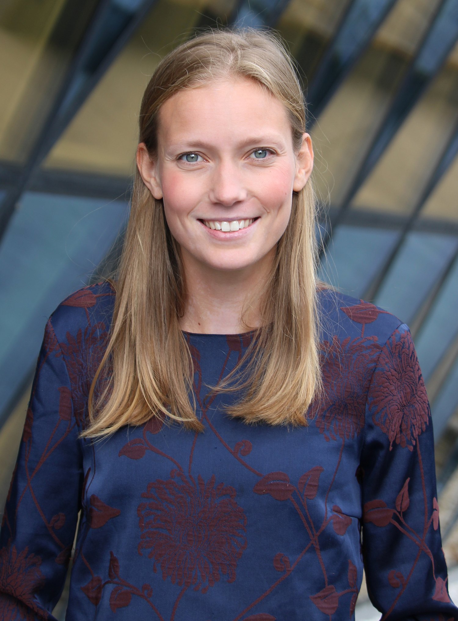 Kvinna med blont hår klädd i en blå topp står framför en byggnad med glasfasad.