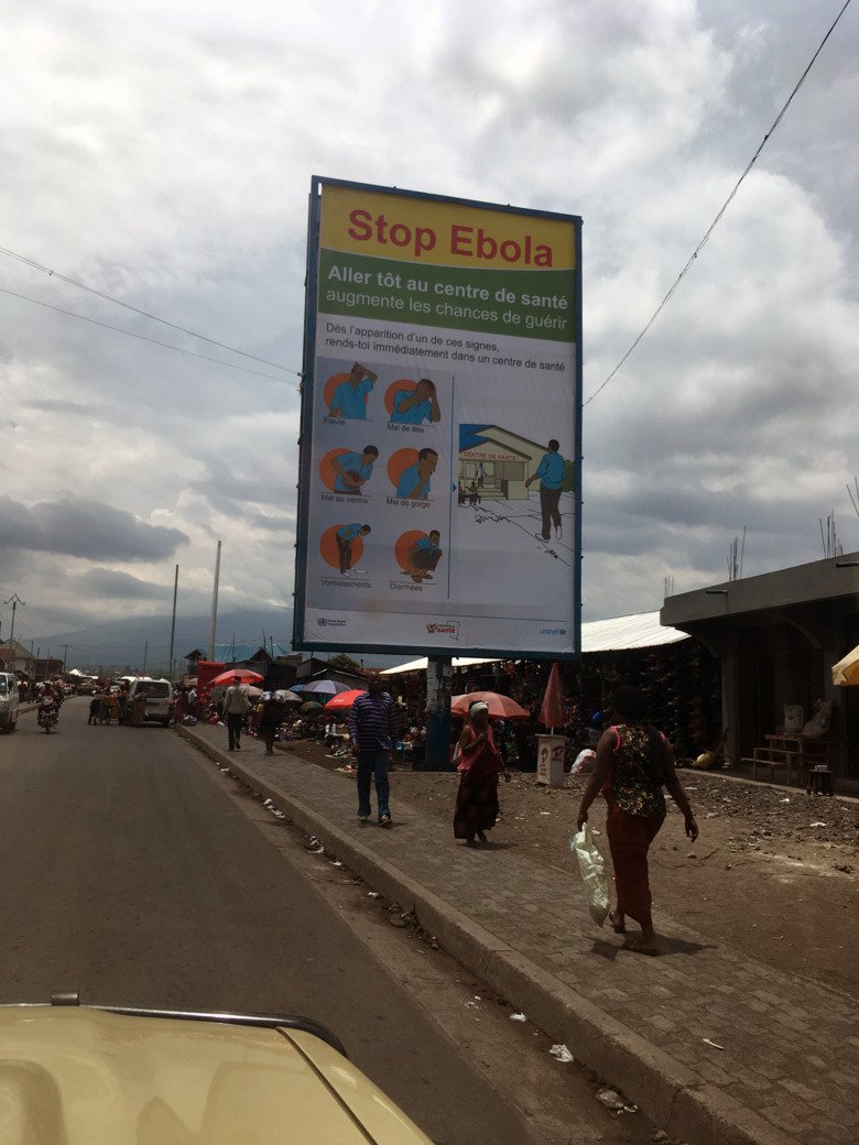 Skylt med information om Ebola längst en gata med bilar, männiksor och hus i DR Kongo.