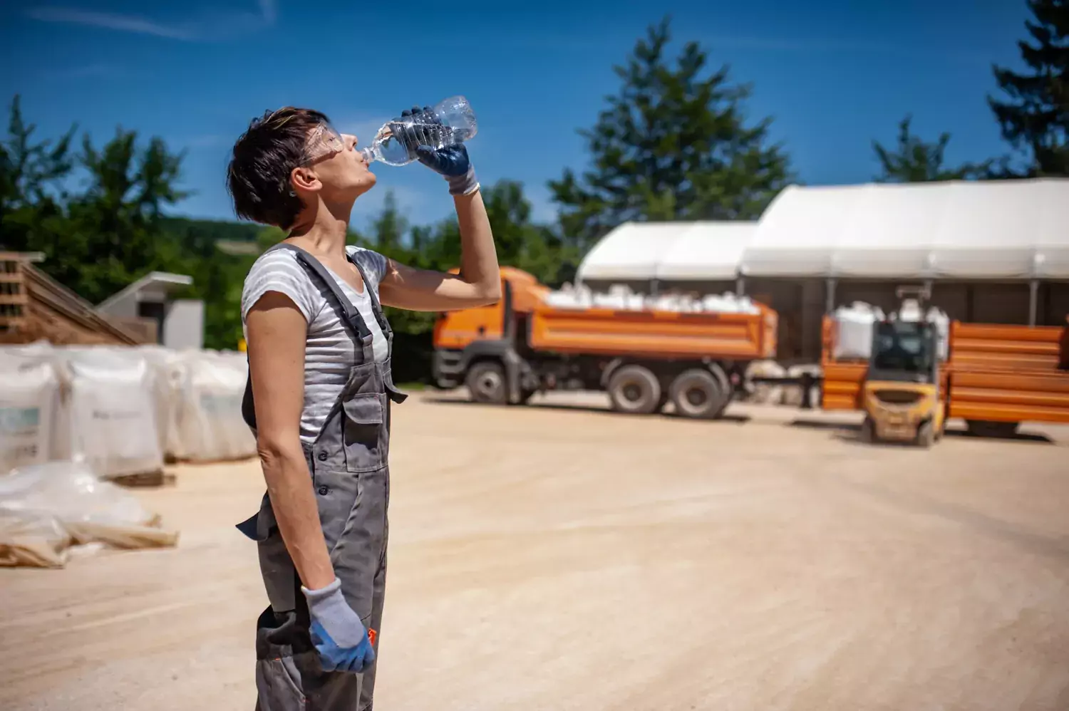 Ung kvinna i arbetskläder dricker vatten ur en flaska framför en dammig väg med lastbilar