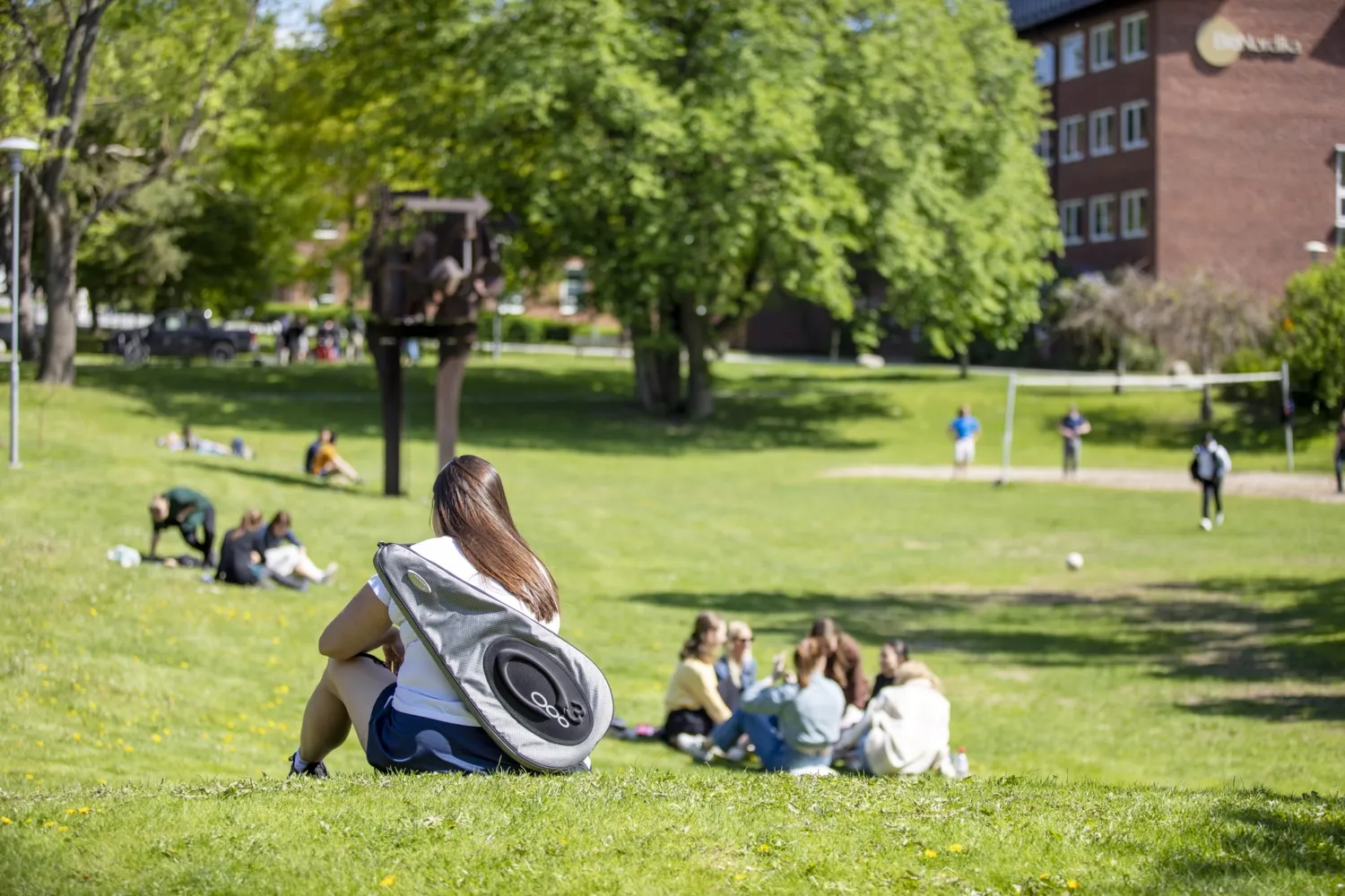 Kvinna med badmintonrackväska på ryggen sitter på gräsmatta på campus Solna, med personer i bakgrunden.