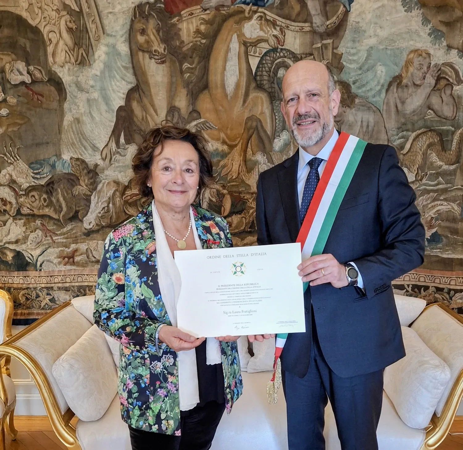 Professor Laura Fratiglioni, NVS, tilldelas utmärkelsen Riddare av italienska Stjärnorden av Italiens ambassadör i Stockholm Michele Pala. De står framför en vacker gobeläng och en guldförgylld soffa.