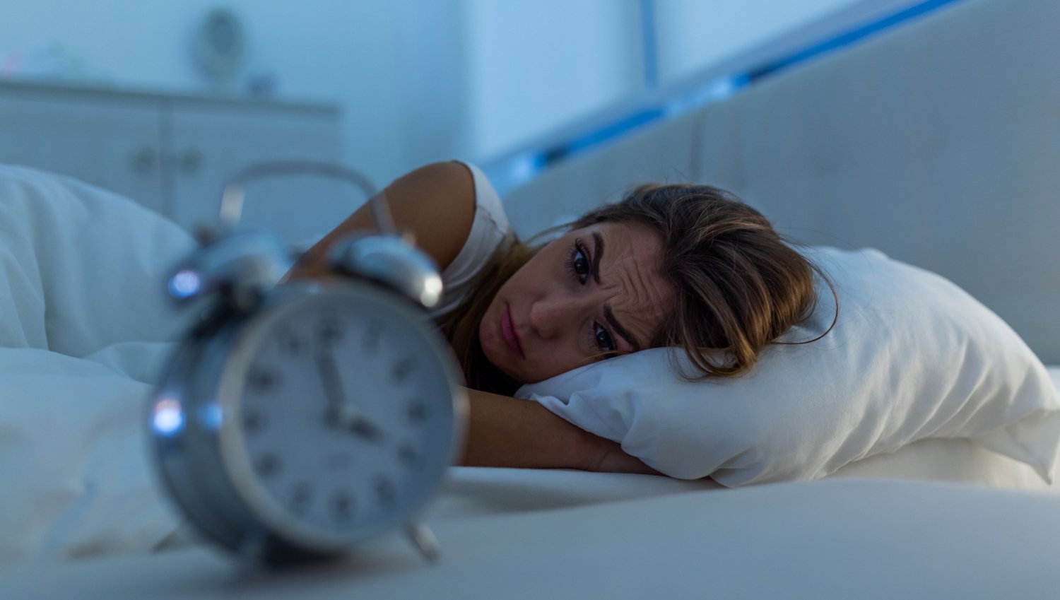 Kvinna som ligger i sängen och rynkar pannan. I förgrunden en klocka med visarna som pekar på klockan fyra. Det är morgonljus i fönstret ovanför sängen.