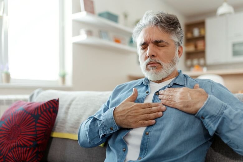 En man i övre medelålder med vitt skägg sitter i soffan och håller sig oroligt om hjärtat