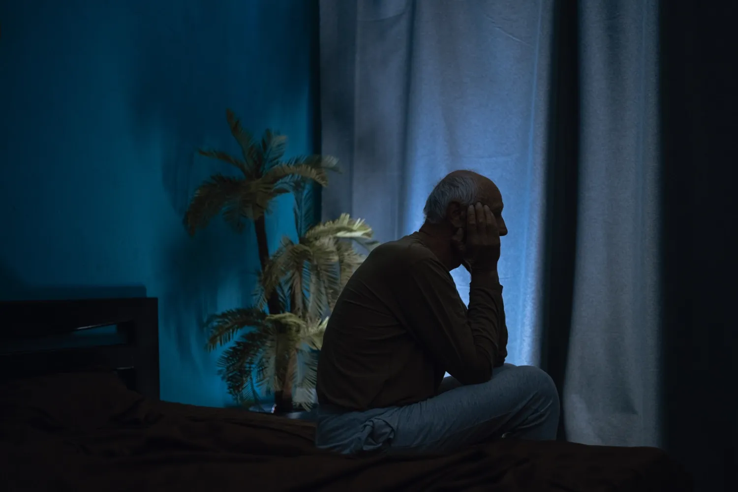 Deprimerad man sitter på sängkanten i ett mörkt rum, med gardinerna fördragna.