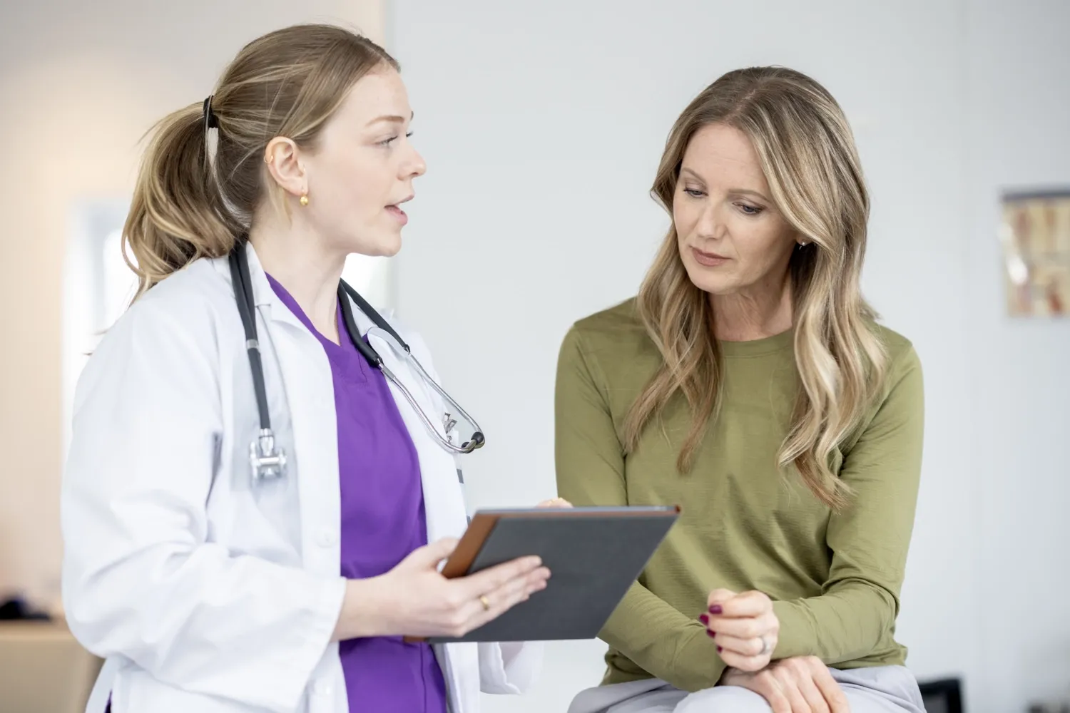 Kvinnlig läkare med lila tröja, vit rock och stetoskop runt halsen pratar med kvinnlig patient i grön tröja..