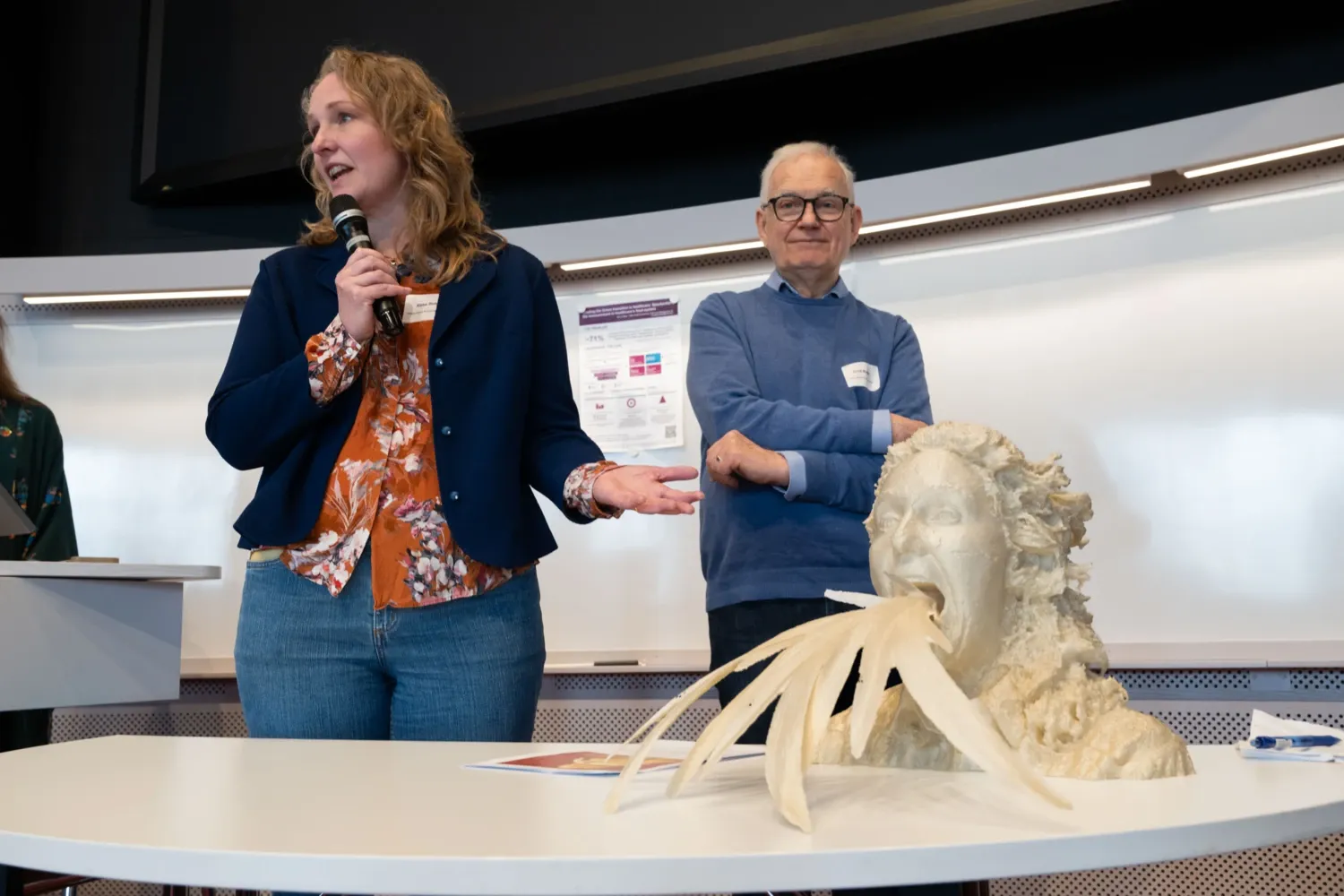 Ett tredimensionellt konstverk föreställande ett huvud med växtliknande former som kommer ut ur munnen står på ett bord i en föreläsningssal. I bakgrunden syns deltagare vid KI:s hållbarhetsdag.