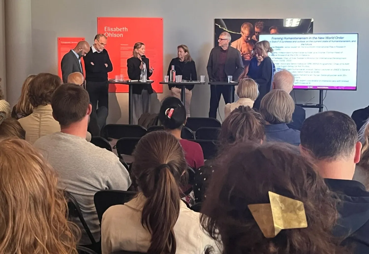 Sex personer står bakom ett ståbord, framför sitter en publik.