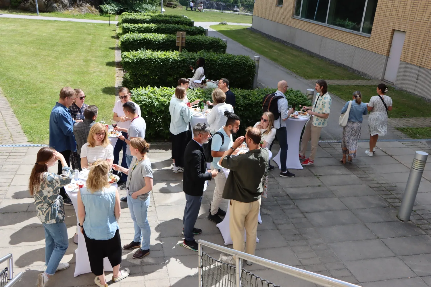 NVS sommarlunch 2024. Folk står ute och äter vid runda ståbord med vita dukar, på baksidan av ANA 23.