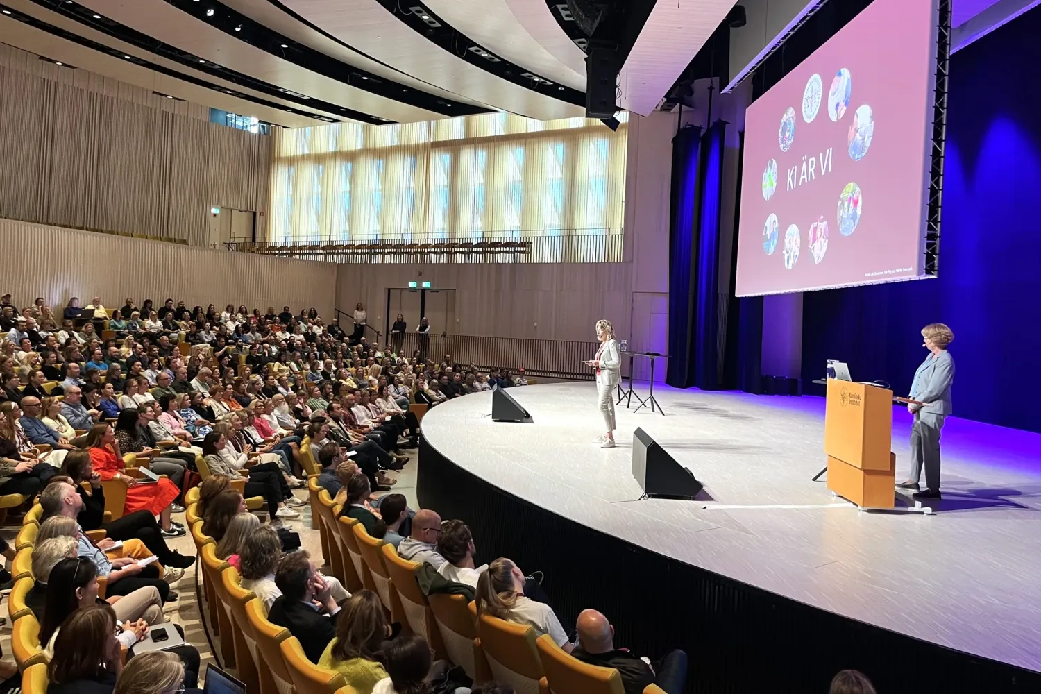 Rektor i beige kostym står på scenen i Aula Medica och talar framför många åhörare i de gula bänkarna framför. I bakgrunden syns en bild med texten "KI är vi".