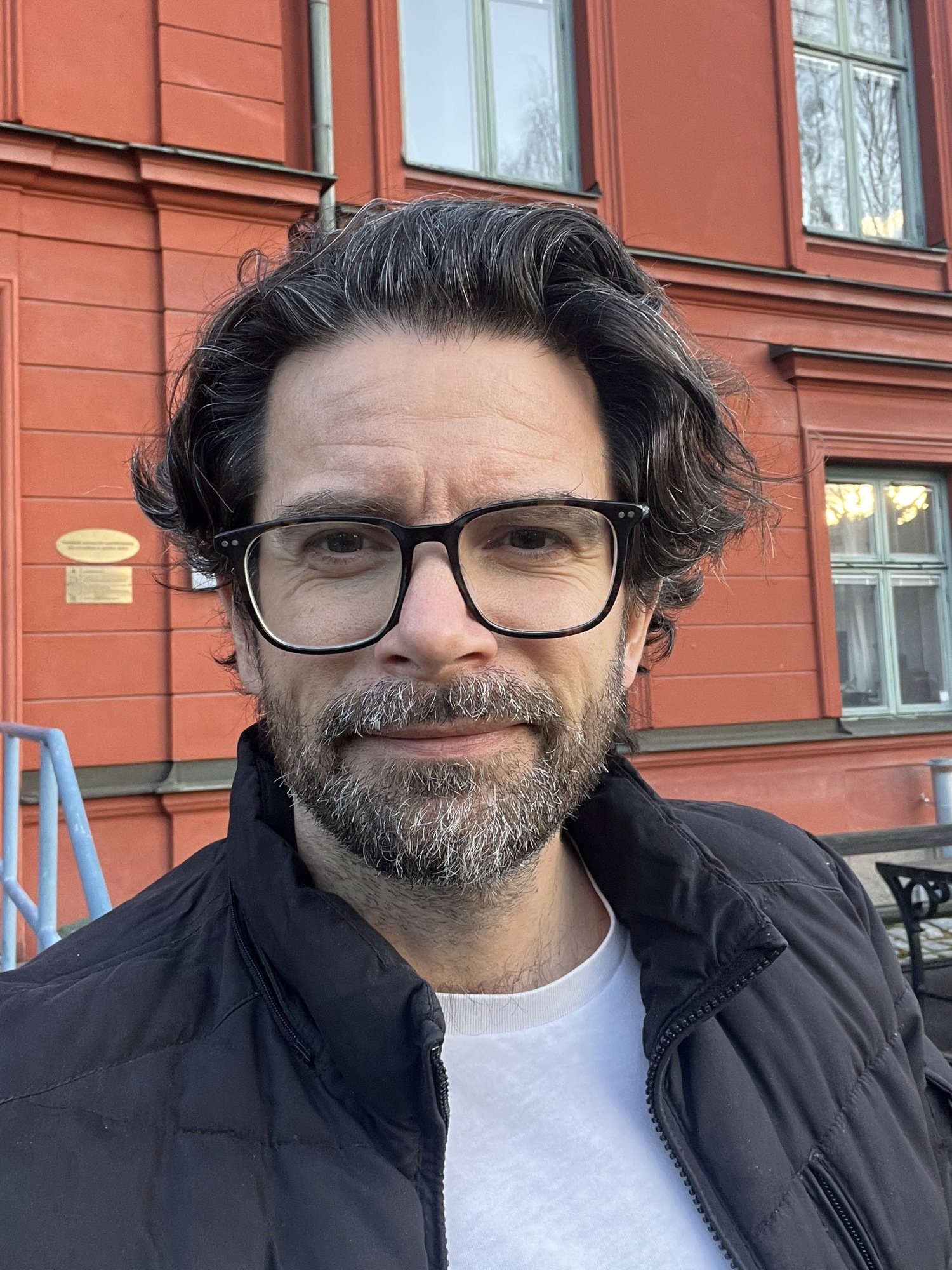 Ansiktporträtt av Gergö Hadlaczky som står utomhus framför en röd byggnad. Han har en glasögon, en vit tshirt och en svart jacka.