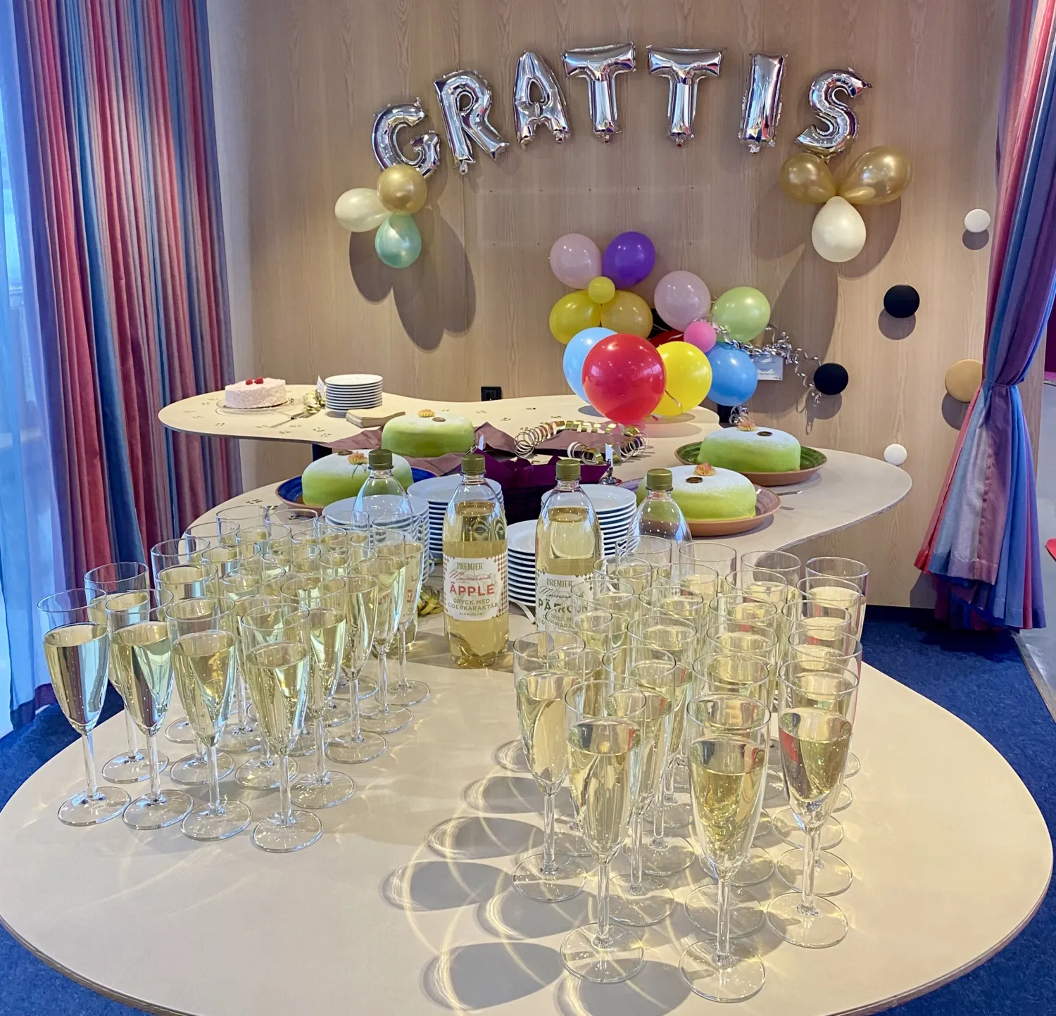 Ciderglas på bord i förgrunden, princesstårtor bakom dem på bord. Grattisballonger och färgglada balonger i bakgrunden.