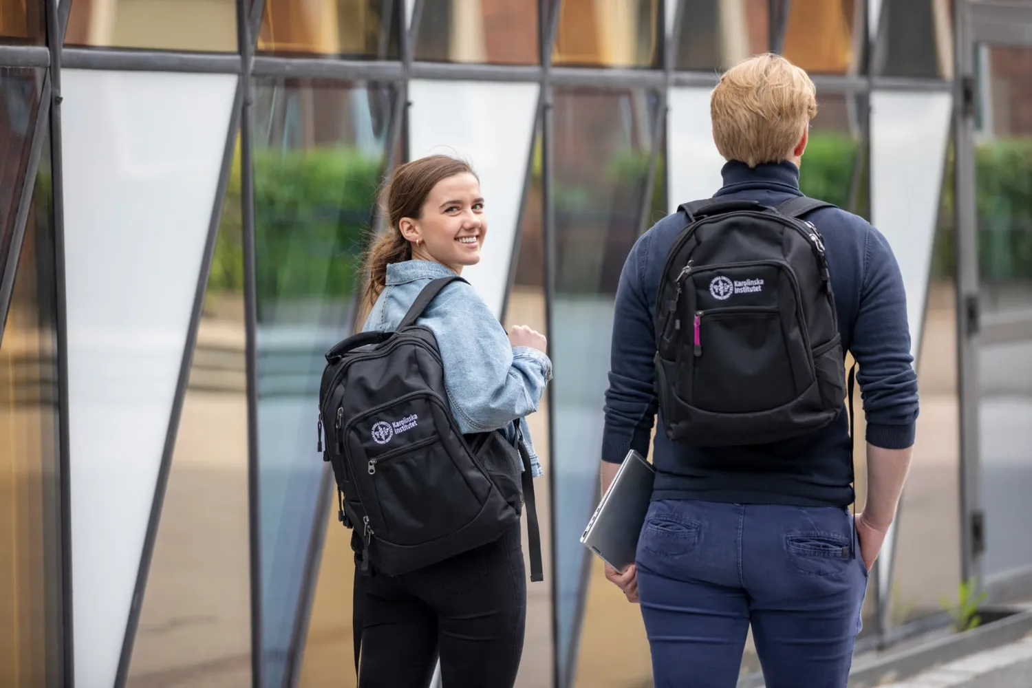 Två studenter utanför Aula Medica med KI ryggsäckar. Den ena vänder sig om och tittar leende mot kameran. || Två studenter går framför Aula Medica med KI-ryggsäck på ryggarna.