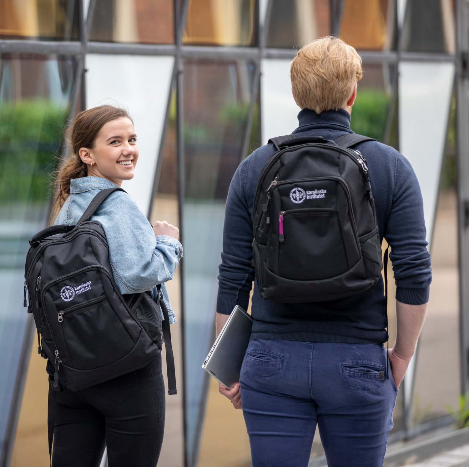 Två studenter utanför Aula Medica med KI ryggsäckar. Den ena vänder sig om och tittar leende mot kameran. || Två studenter går framför Aula Medica med KI-ryggsäck på ryggarna.