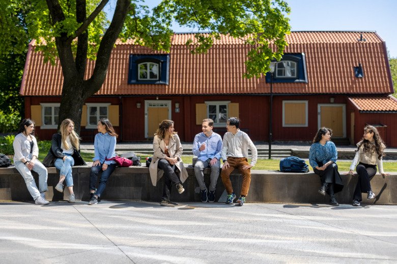Flera glada studenter sitter på en mur framför ett rött hus på ett soligt campus Solna.