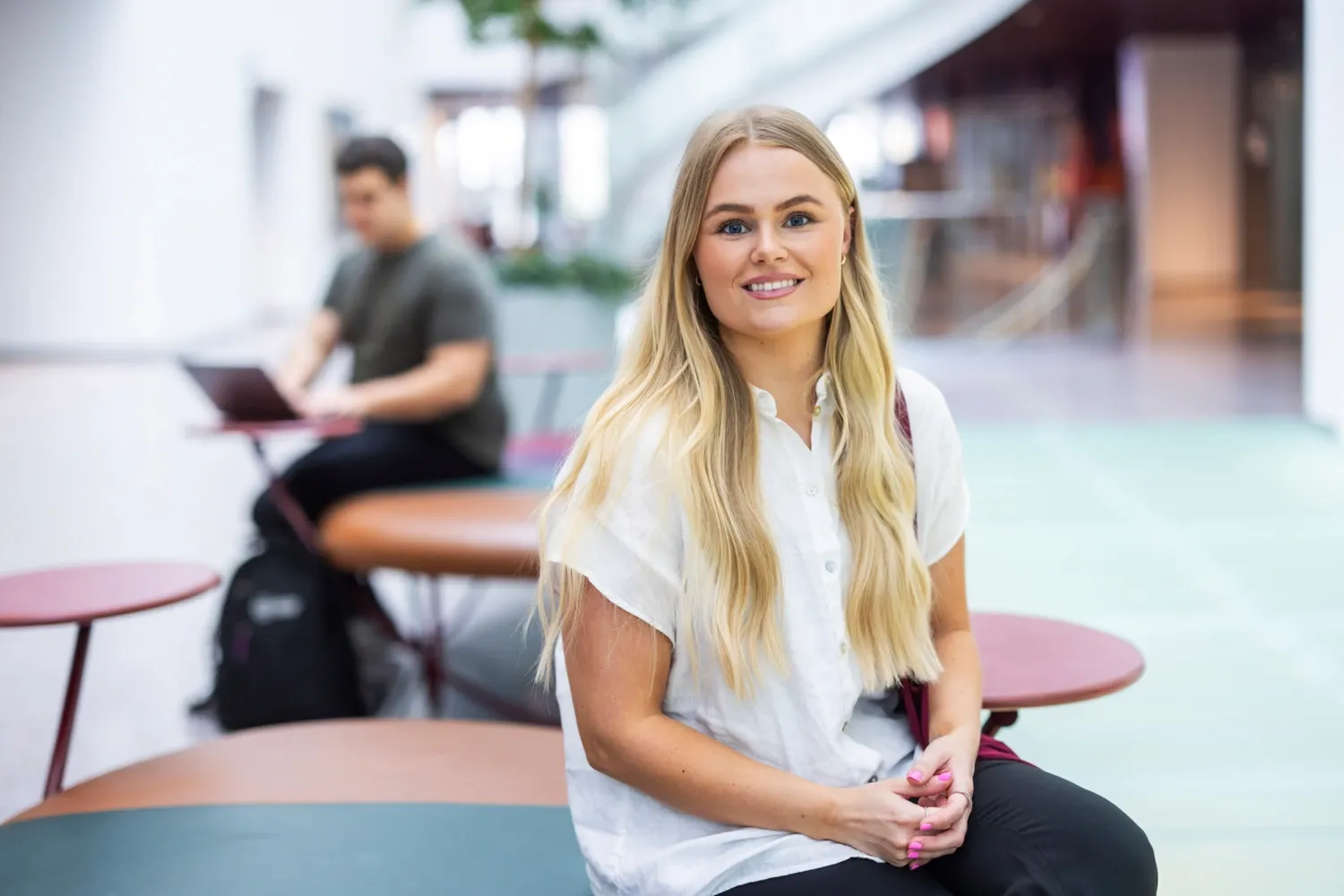 ung kvinna i långt ljust hår sitter på en bänk innomhus på ett ljust och modernt campus.