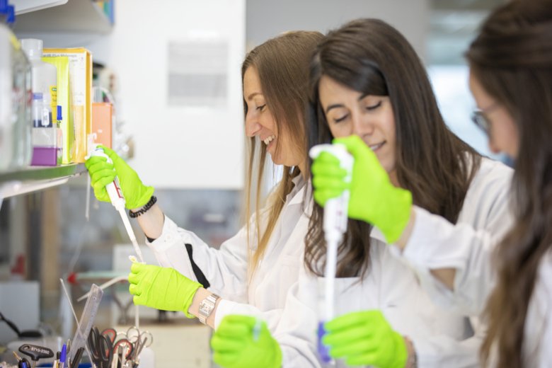 Tre kvinnliga forskare står med provrör och pipetter i labbmiljö.