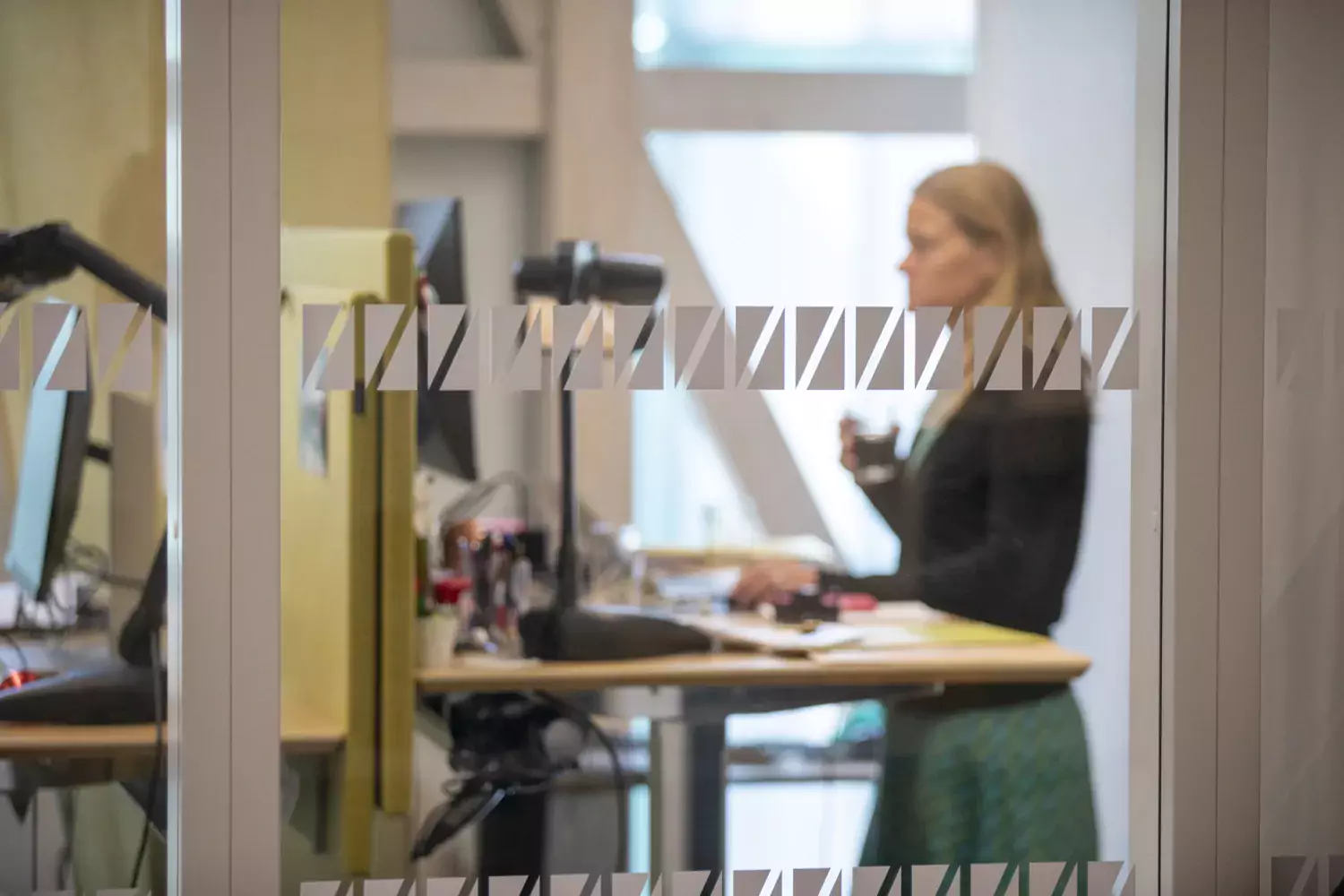 I förgrunnden: glasvägg med motiv. I bakgrunden: Kvinna står vid ett skrivbord i öppet kontorslandskap.