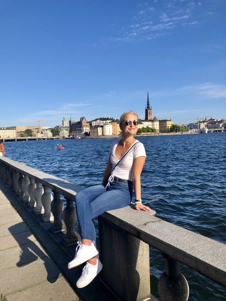 Kvinnlig student som sitter på en mur framför vattnet och i bakgrunden ser man Stockholm och vädret är fint.