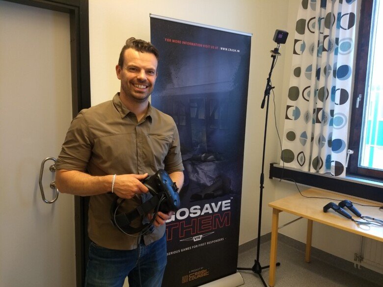 En man visar upp VR utrustning bredvid en affisch som visar programmet 'Go save them VR'