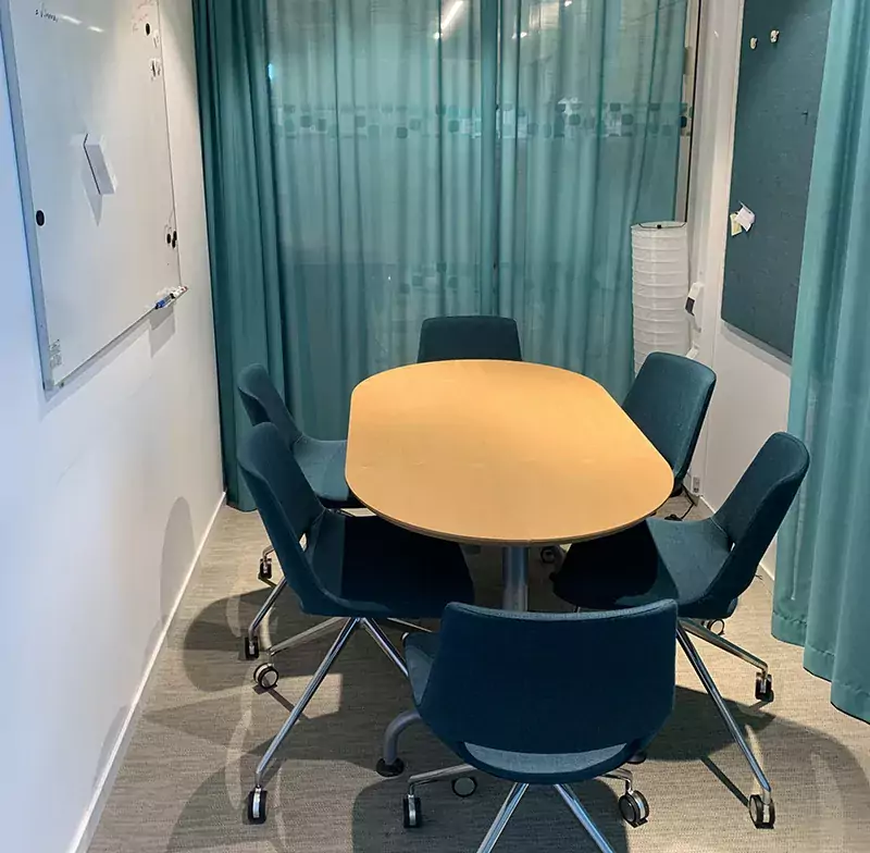 Bild av ett mötesrum med sex blå stolar och ett träbord. Gröna tunna gardiner omger väggarna.