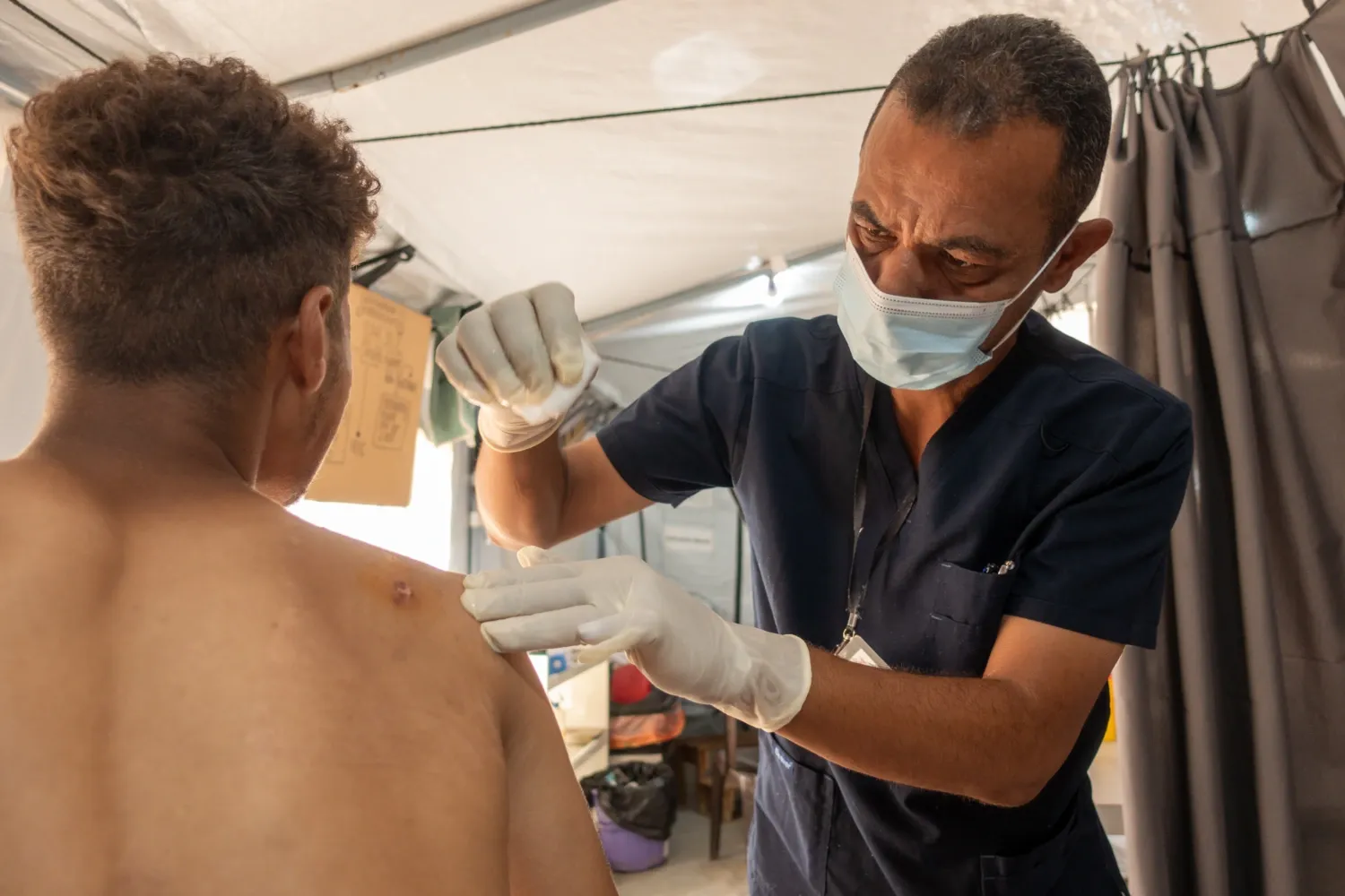 En man sitter i bar överkropp med ryggen mot kameran. Han har ett sår i axeln. En sjukvårdare med munskydd och plasthandskar rengör såret. Bakgrunden syns att de står i ett sjukvårdstält.