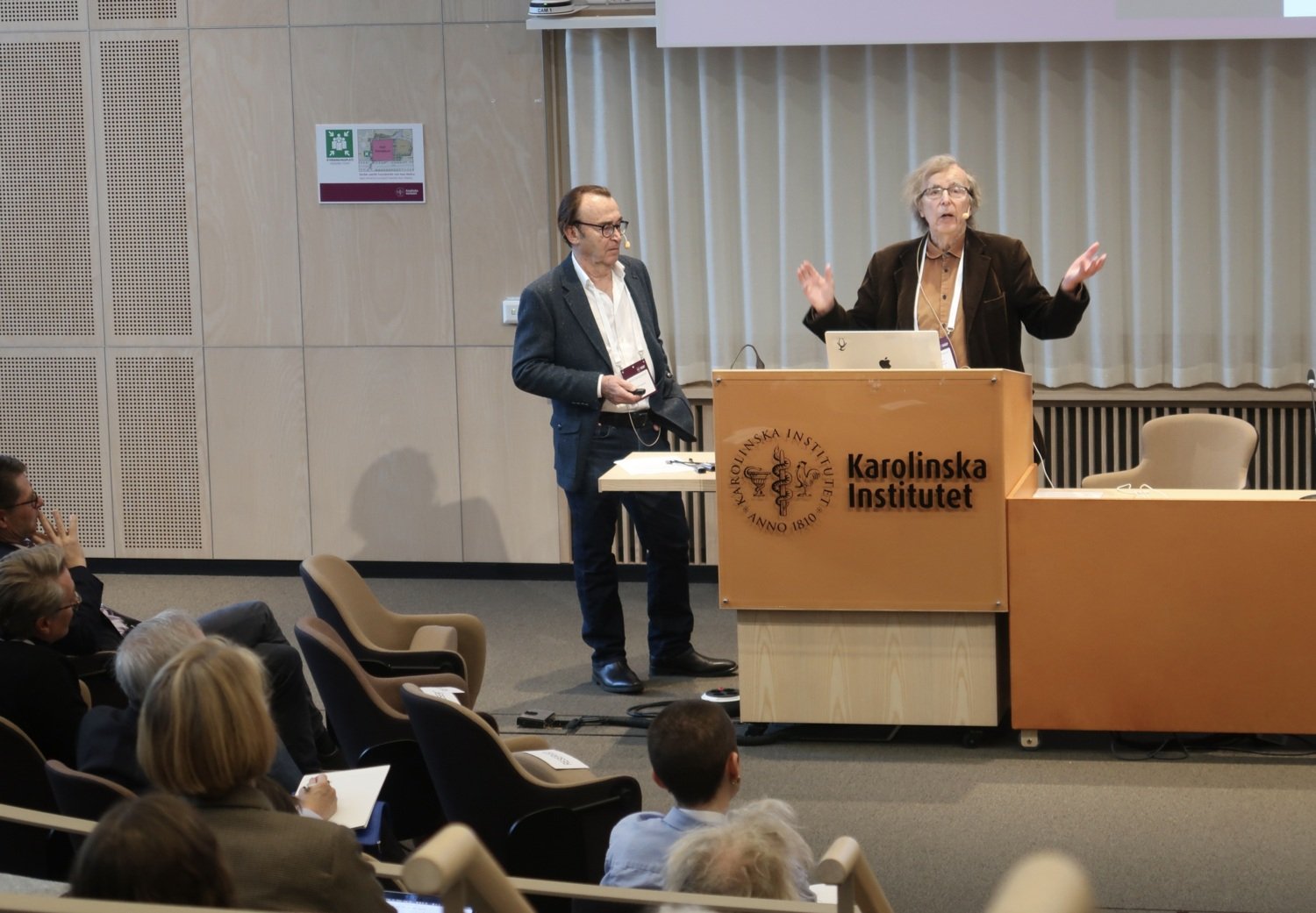 Två män står vid pulpeten längst fram i en föreläsningssal och talar.