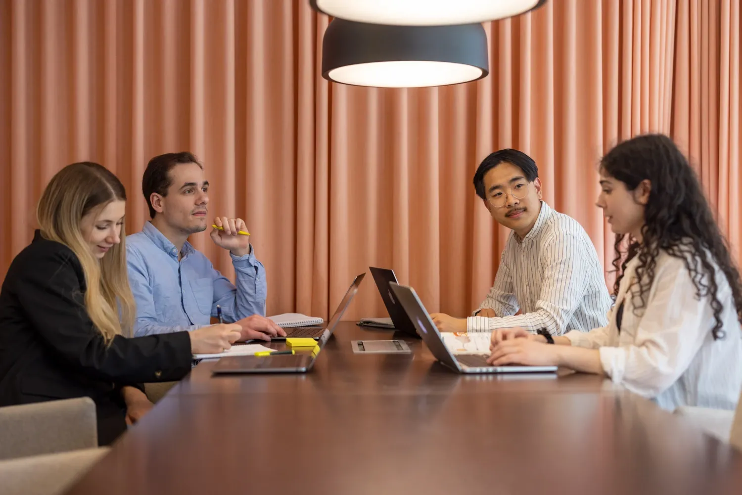 Fyra personer sitter vid ett bord med datorer och anteckningsblock framför sig.