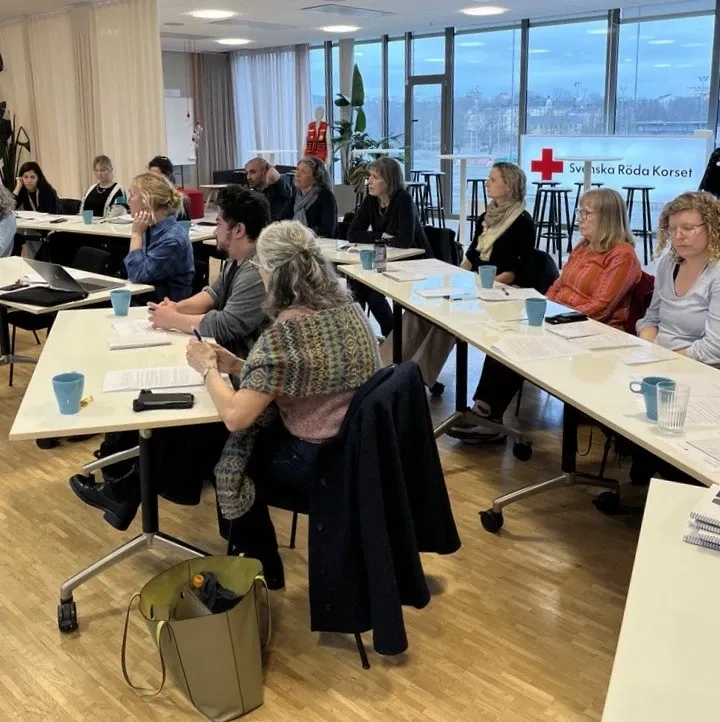 Personer sitter i en undervisningssal, i bakgrunden syns en skylt med texten Röda korset och några fönster med utsikt över havet