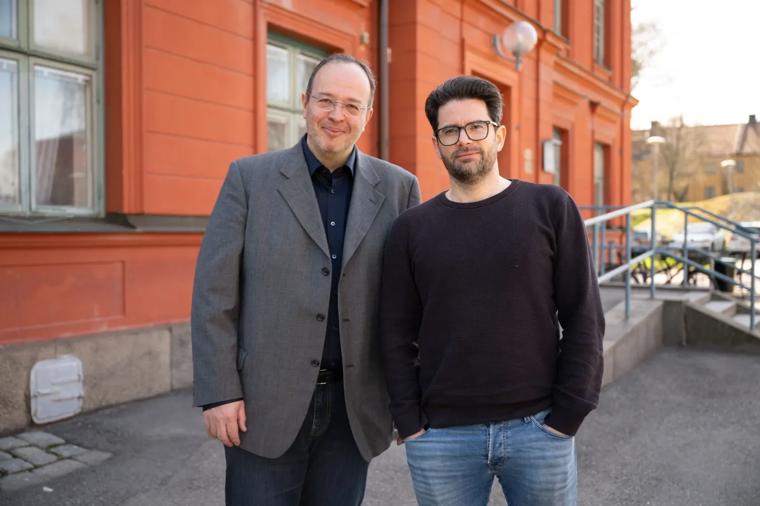 Vladimir Carli, iklädd en grå kavaj, och Gergö Hadlaczky, iklädd en svart tröja, står fram för en röd byggnad.