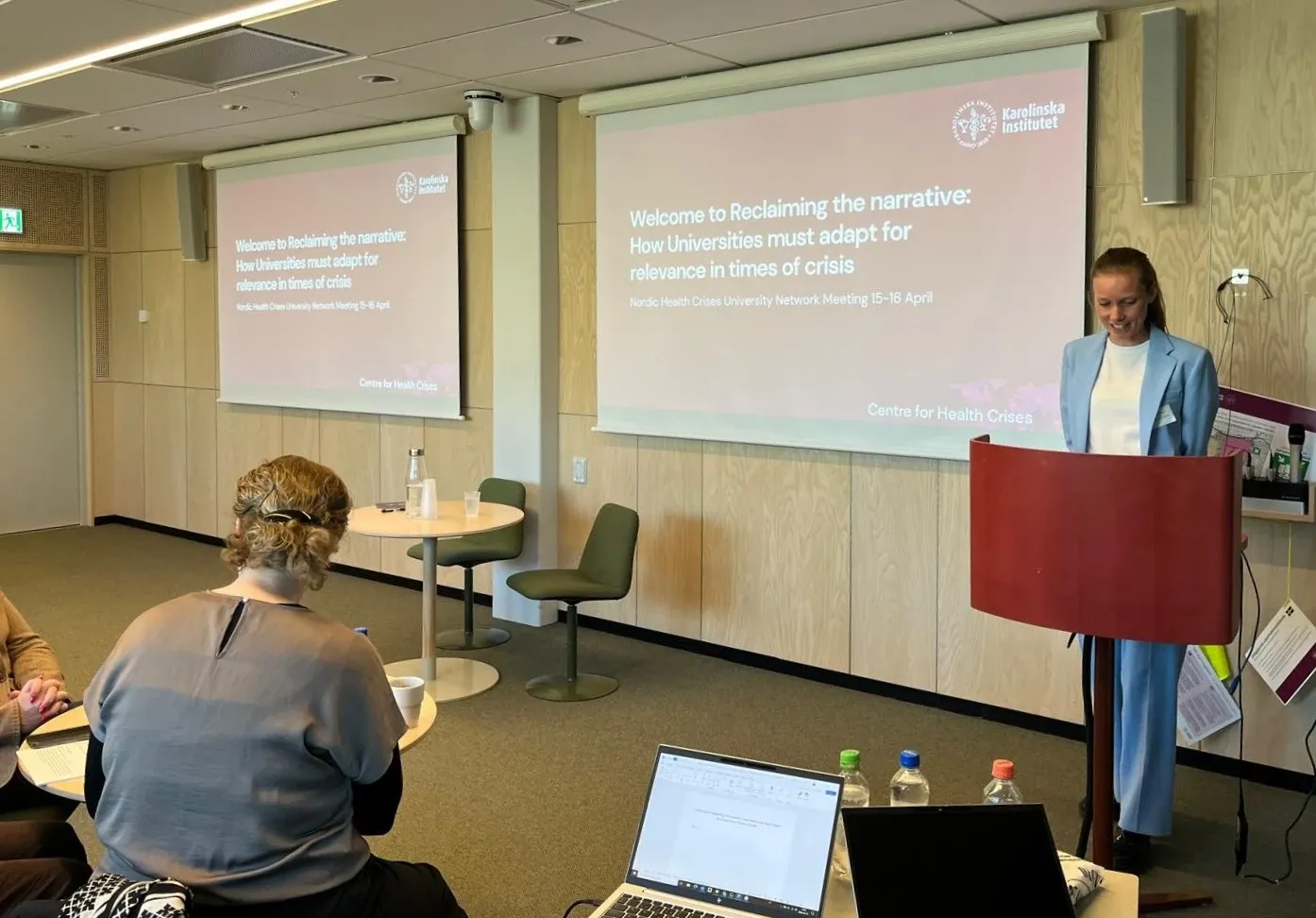 En kvinna står framför en projektorduk där det står Reclaiming the narrative: How Universities Must Adapt for Relevance in Times of Crisis och vi ser ryggen på en annan kvinna som sitter framför