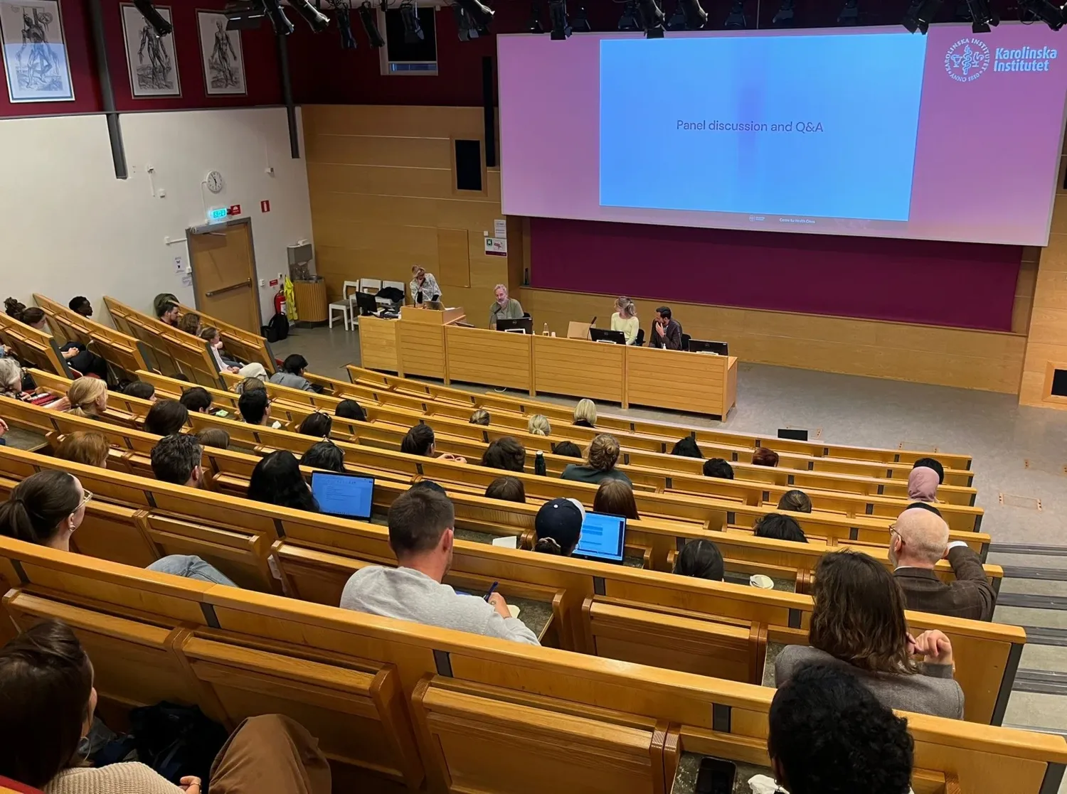 En föreläsningssal med människor som tittar på fyra personer och en powerpoint presentation