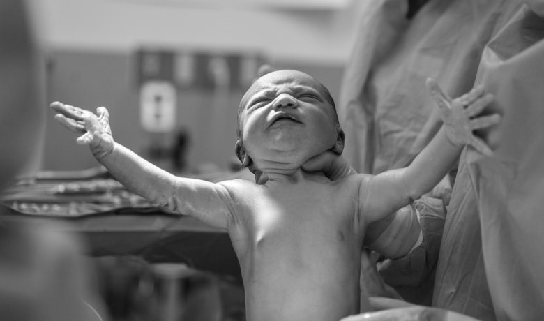 Två händer håller upp ett nyförlöst spädbarn.