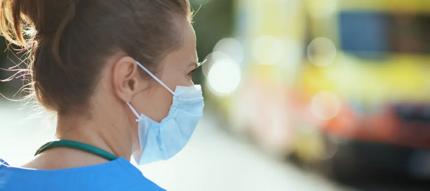 En kvinna med munskydd som vänder sig åt sidan; i bakgrunden skymtar en ambulans