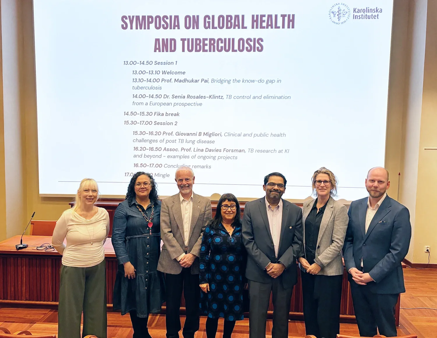 sju personer som står i en aula. Bakom visas en stor bildskärm.