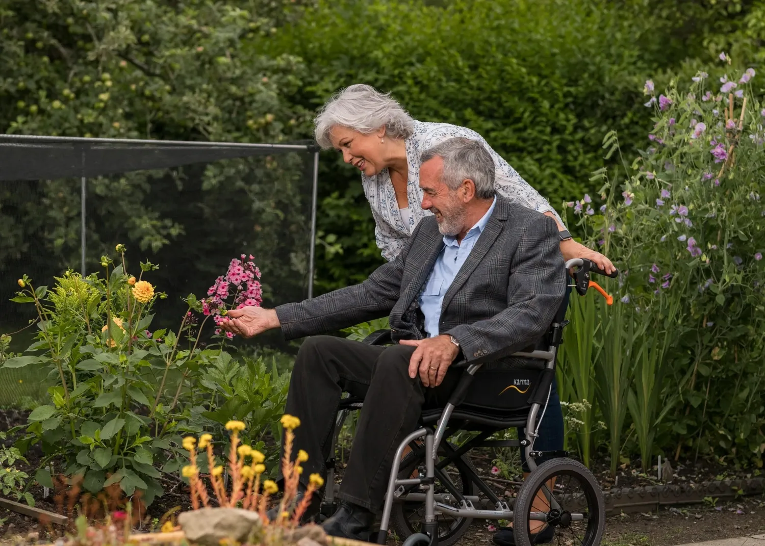 Kvinna står bakom man i rullstol i en trädgård, tittar på blommor