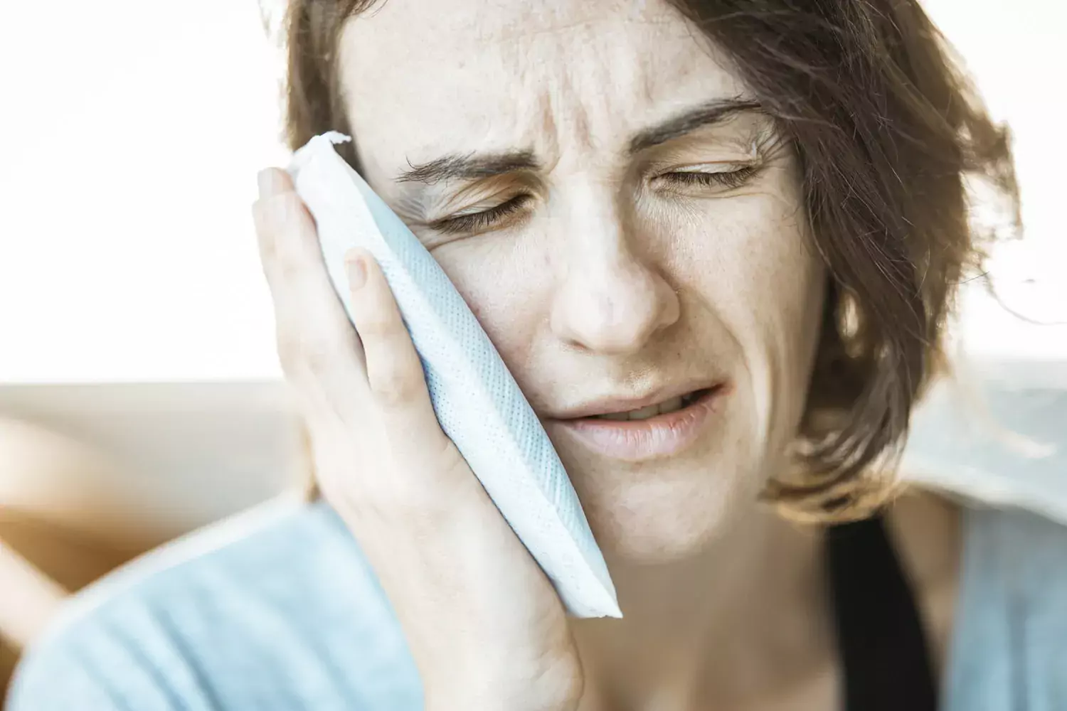 Bilden visar en kvinna som har smärta i käken. Hon håller en kylklamp mot sin högra kind och blundar av smärtan.