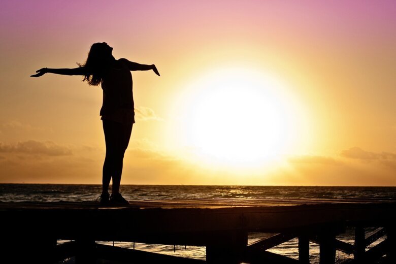 Kvinna står på brygga i solnedgången med armarna utsträckta.