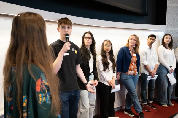 Studenter står på rad i en föreläsningssal. En person talar i mikrofon medan andra deltagare håller papper i händerna.