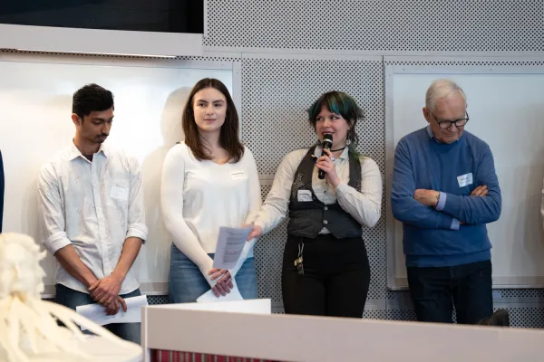 Studenter står på rad i en föreläsningssal. En person talar i mikrofon medan andra deltagare håller papper.