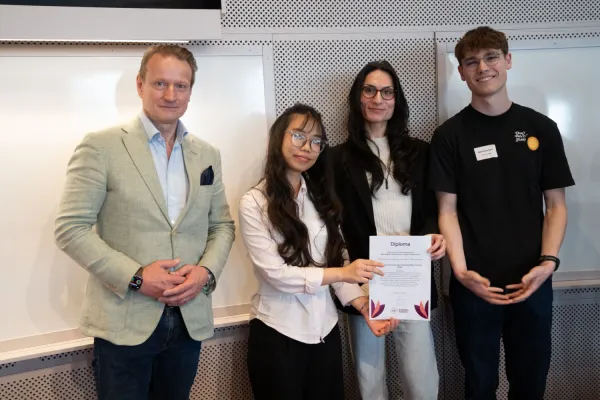 Fyra personer står tillsammans i en föreläsningssal. En person håller upp ett diplom medan de andra står bredvid.