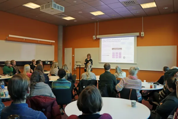 Kristina Dalin Eriksson presenterar sin poster i en föreläsningssal med publik som sitter vid runda bord i förgrunden.