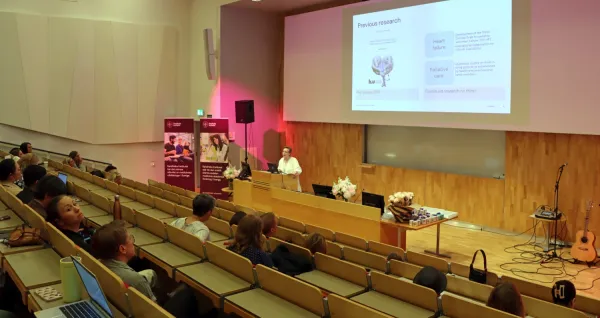 Nana Waldréus, docent i omvårdnad, föreläser om sin forskning vid NVS sommarlunch 2025. Hon står framme vid podiet i H1, med publik i förgrunden.