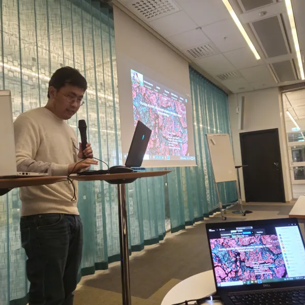 Foto på Yizhe Sun som står längst framme i ett större mötesrum med mikrofon i handen.